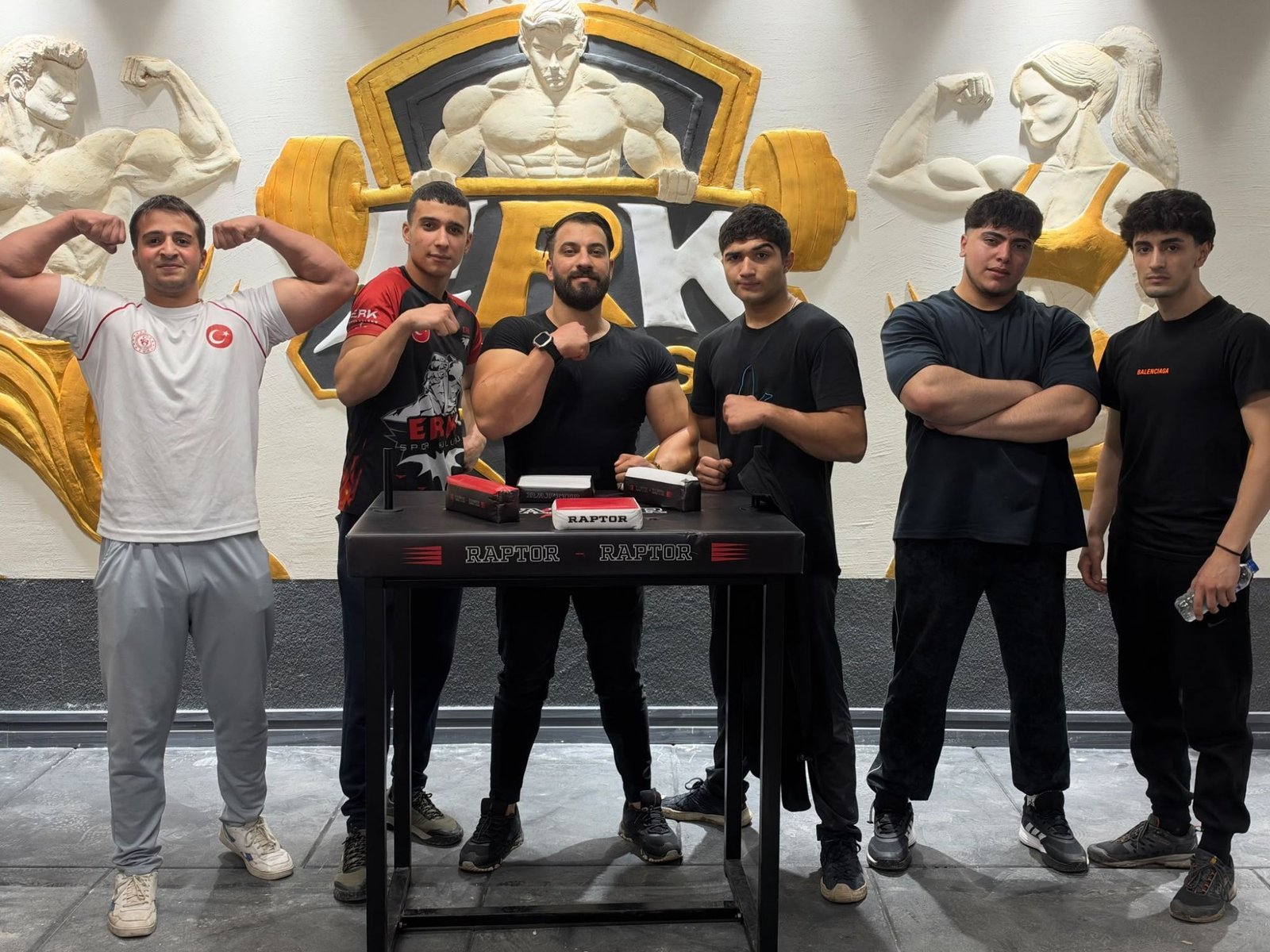 ERK Fitness Clup salonunda, kabartma logolu duvarın önünde bilek güreşi masasının çevresindeki ERK sporcuları